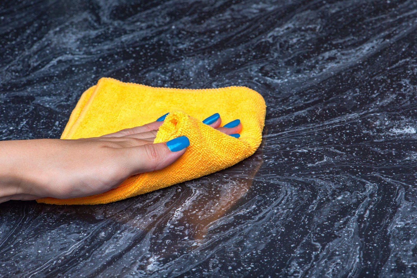 A woman with blue nails cleans a marble counter using a yellow cloth